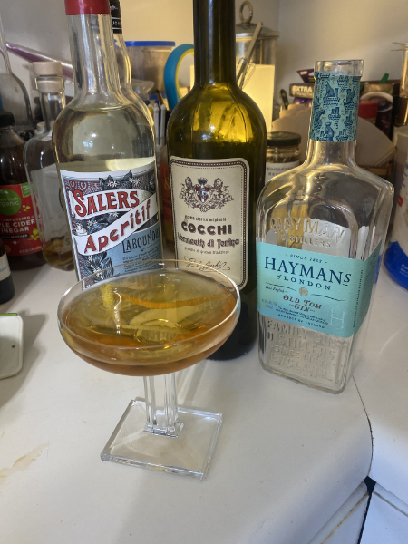 a cocktail coupe with a drink in it, backed by the bottles of the ingredients used: Salers Aperitif, Cocchi di Torino vermouth, and Hayman's Old Tom gin